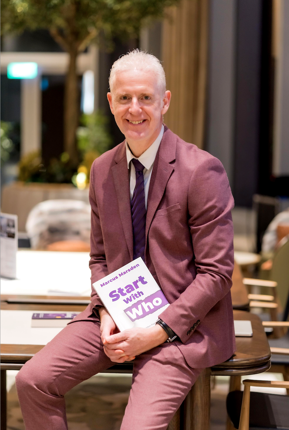 Marcus Marsden in a burgundy suit holding his book Start With Who