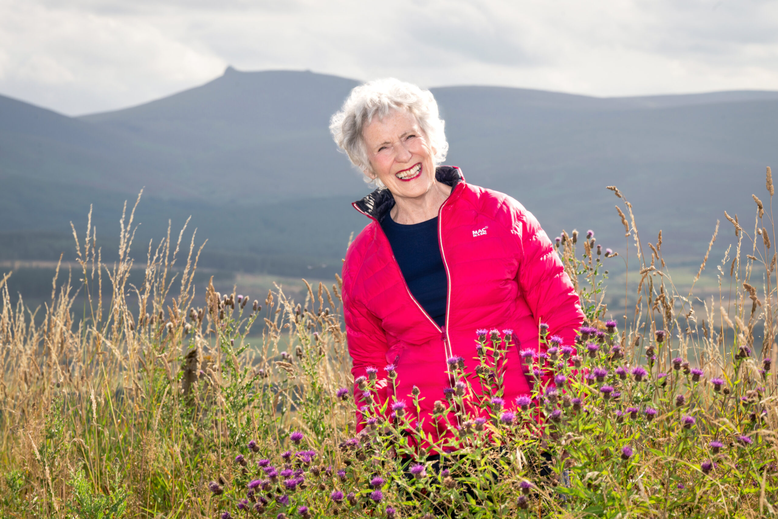 Rosee Elliott laughing in a pink jacket among thistles and long grass, with hills behind her