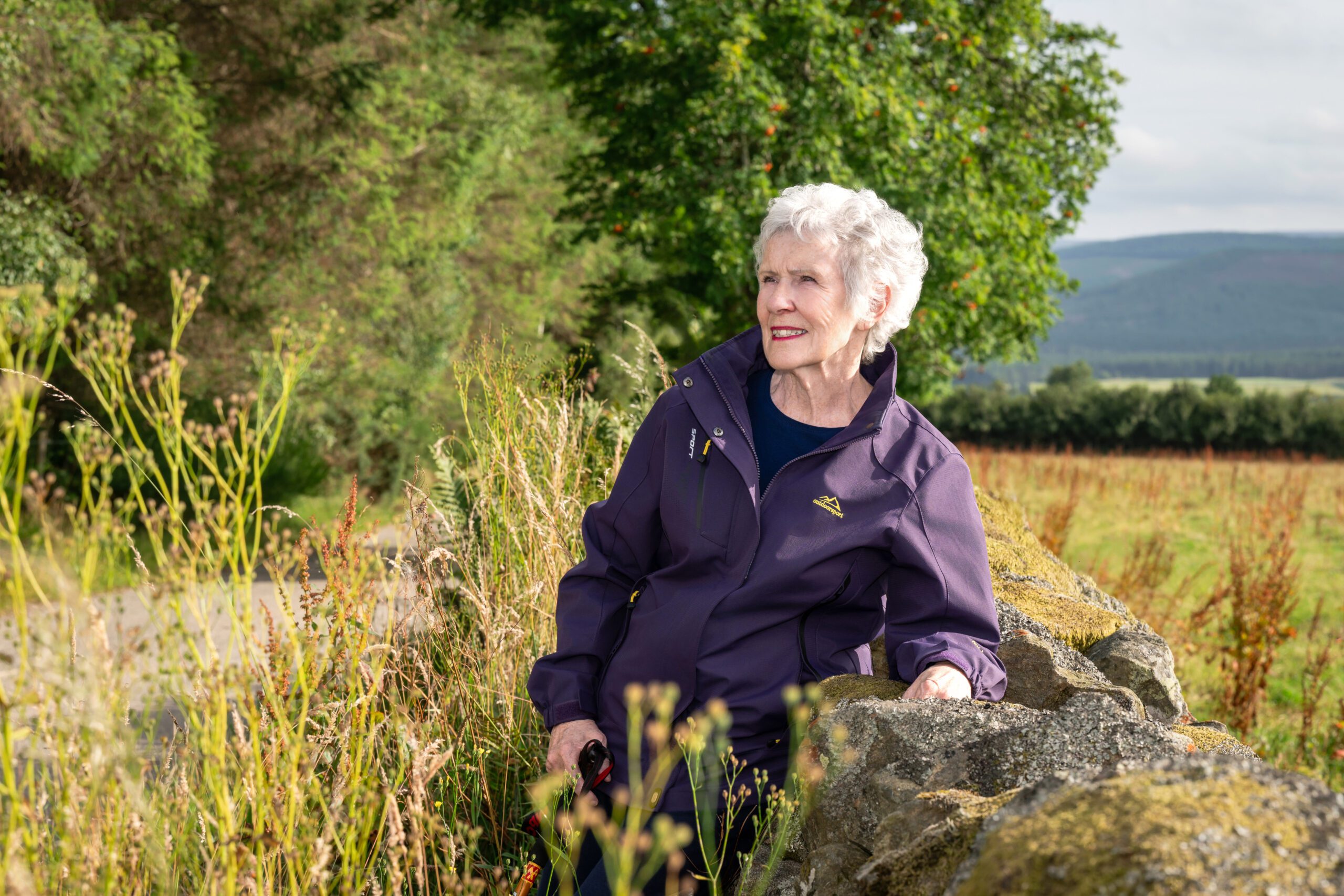 Rosee Elliott in a purple jacket resting against a dry stone wall beside a country road, looking into the distance