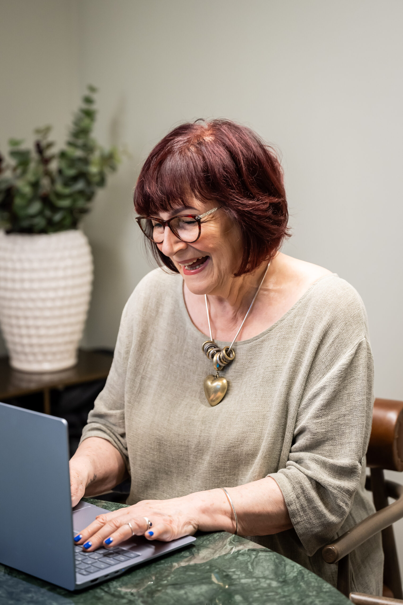 Sharon Eden laughing at a laptop in a beige top and heart pendant, at a green marble table with a plant behind her
