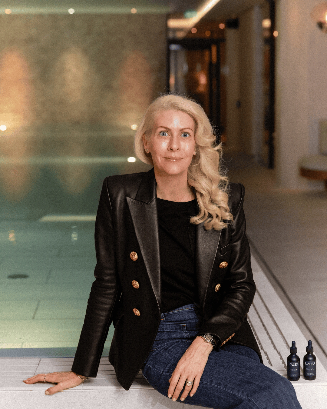 Elin Mendola in a black leather blazer seated beside an indoor pool with two L’AURA bottles