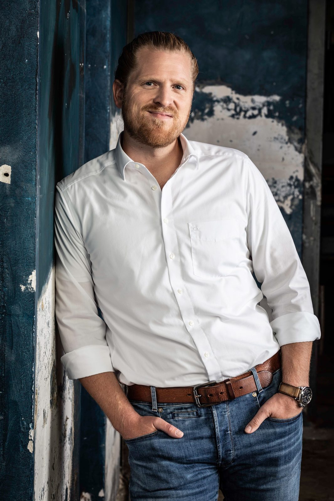 Frederic Lanz standing in a white shirt and jeans, hands in pockets, against a weathered blue wall