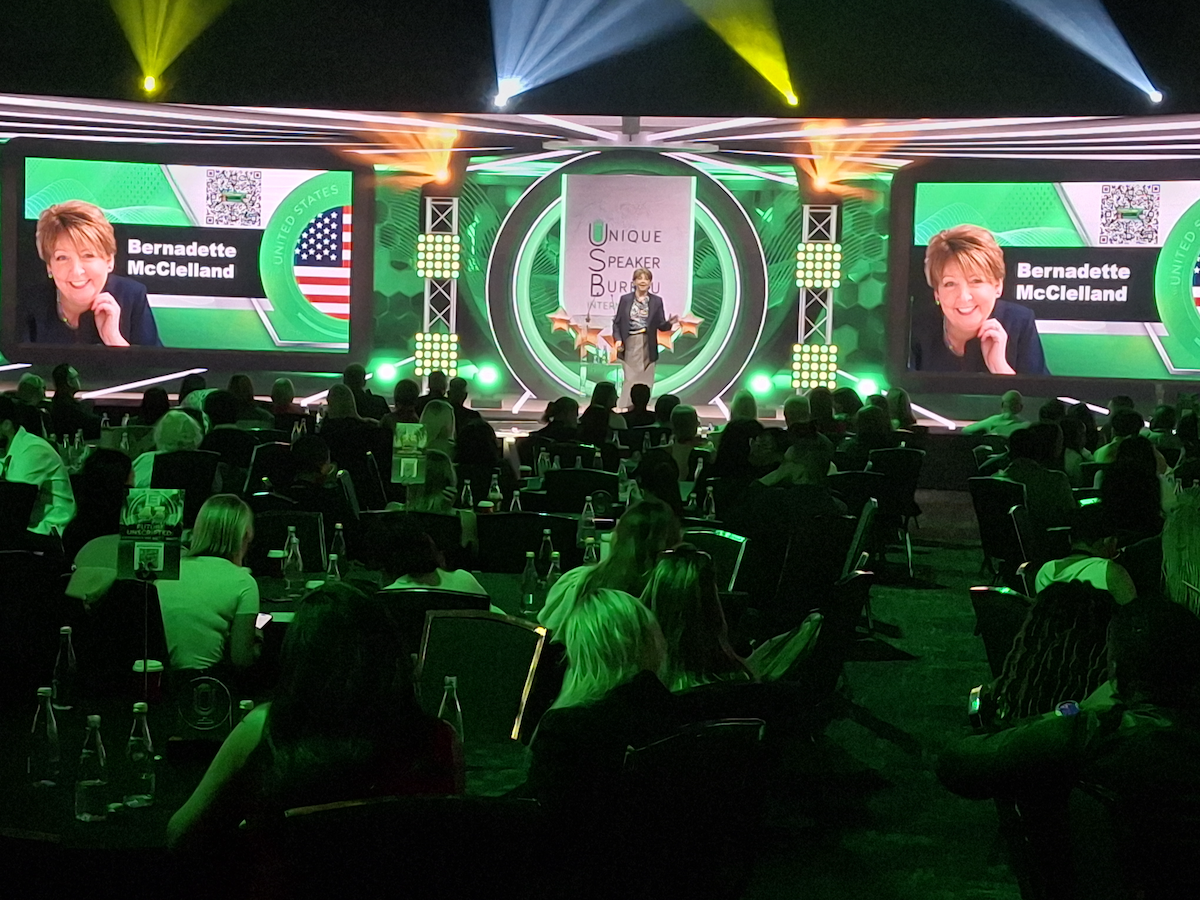 Bernadette McClelland speaking on stage at a Unique Speaker Bureau event, her name and portrait on the screens either side