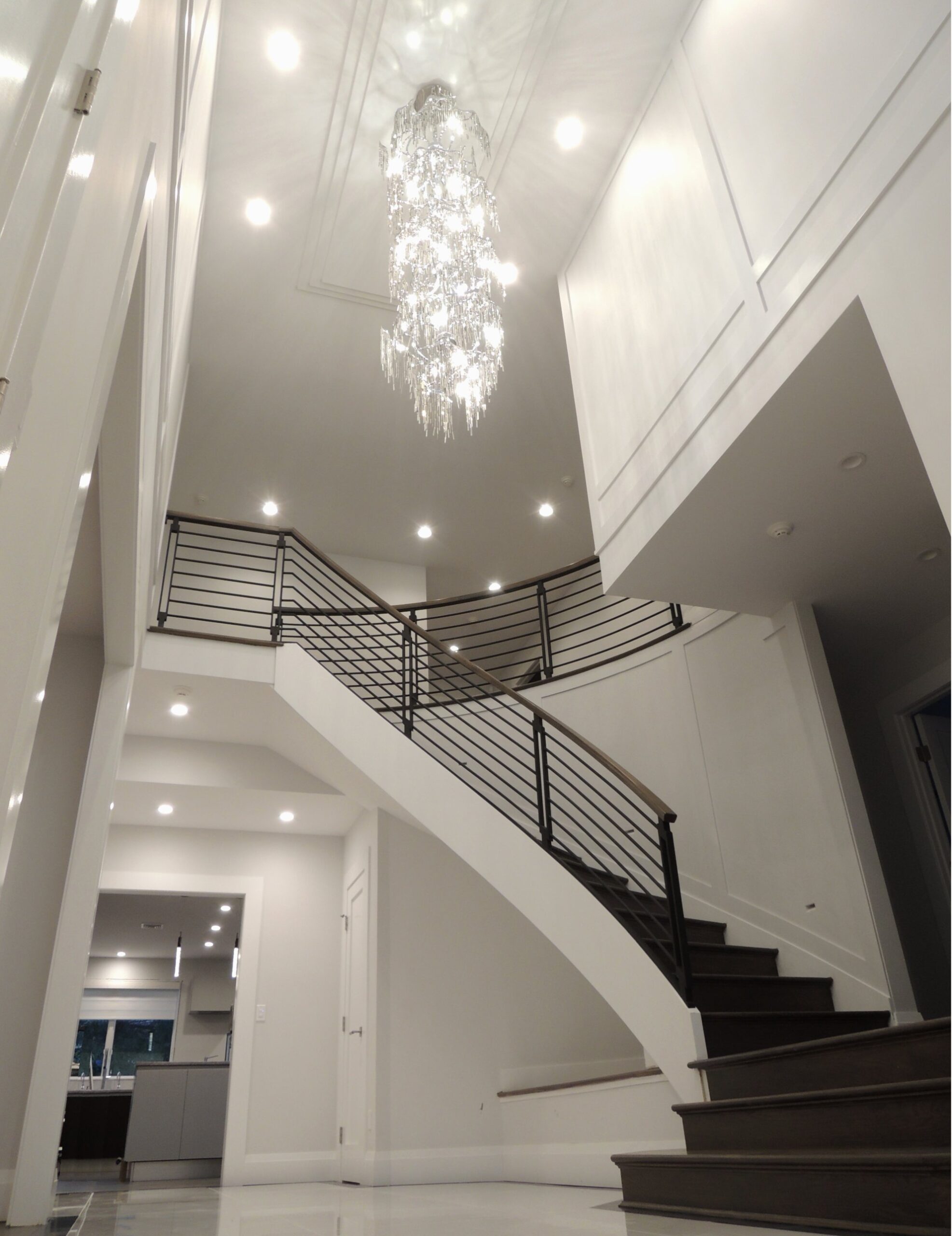 Curved staircase with black railings and a tall crystal chandelier in a white two-storey foyer